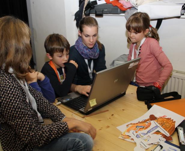 Eine Frau und drei Kinder sitzen an einem Tisch und schauen auf einen Laptop.
