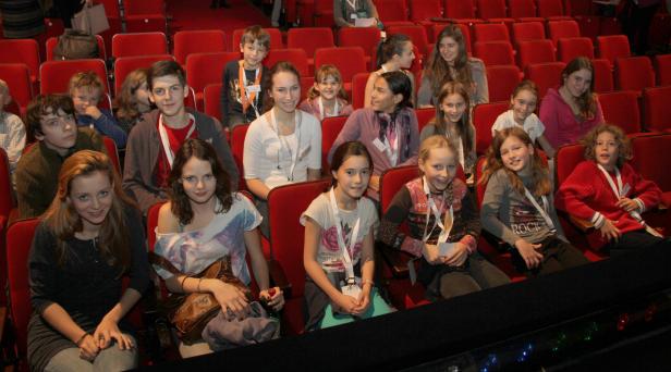 Eine Gruppe von Kindern sitzt in einem Theatersaal auf roten Sitzen.