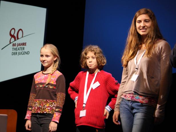 Drei junge Mädchen stehen auf einer Bühne vor dem Logo des Theaters der Jugend.