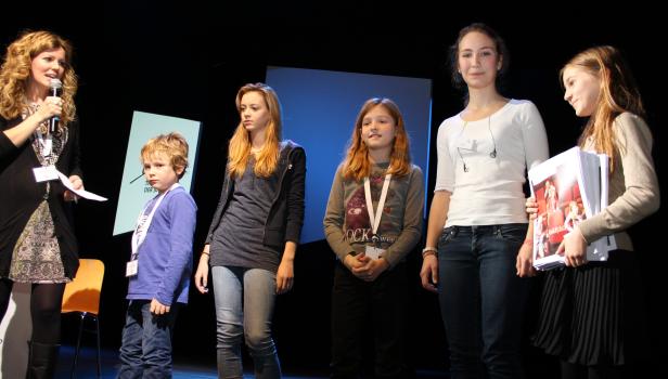 Eine Frau mit Mikrofon steht neben einer Gruppe von Kindern auf einer Bühne.