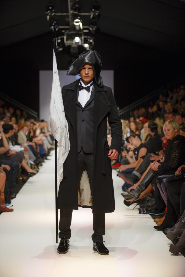 Ein Mann in einem schwarzen Anzug und Mantel mit einem ungewöhnlichen Hut auf dem Laufsteg.