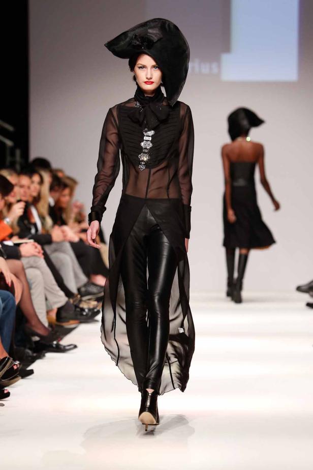 Eine Frau in einem schwarzen Outfit und einem extravaganten Hut auf einem Laufsteg.