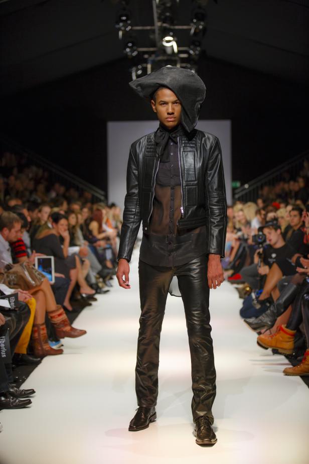 Ein Model in Lederkleidung und einem ungewöhnlichen Hut auf einem Laufsteg.