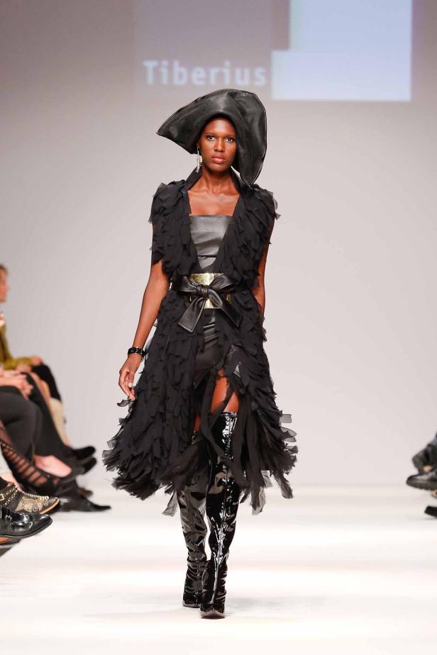 Eine Frau in einem schwarzen Kleid und Hut läuft über einen Laufsteg.