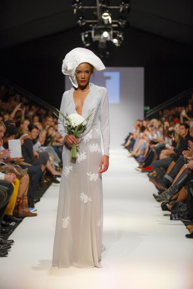 Eine Frau in einem langen Kleid und einem Hut auf einem Laufsteg mit Blumenstrauß.