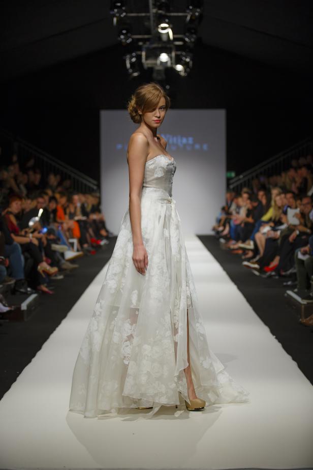 Eine Frau präsentiert ein weißes, trägerloses Brautkleid auf einem Laufsteg.