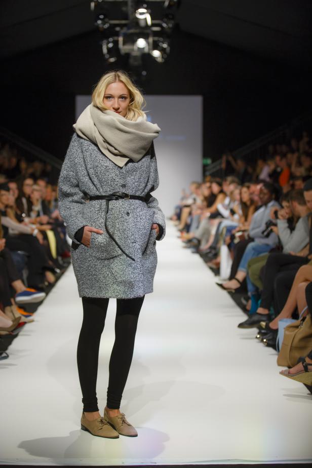 Eine blonde Frau präsentiert einen grauen Mantel und einen beigen Schal auf dem Laufsteg.