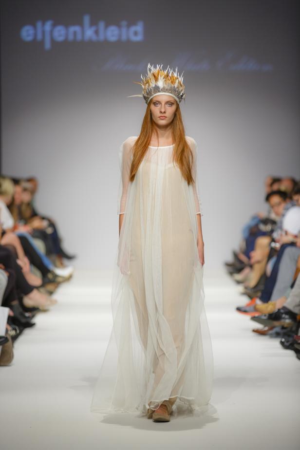 Eine Frau mit Federschmuck präsentiert ein langes, transparentes Kleid von Elfenkleid.