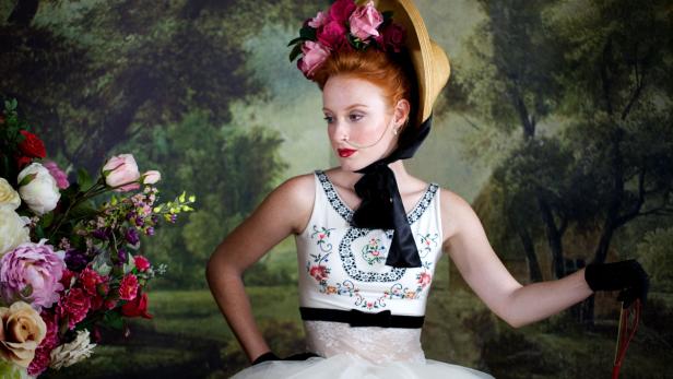 Eine Frau mit Hut und Blumen posiert vor einem gemalten Hintergrund.