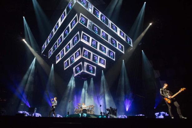 Eine Band tritt auf einer Bühne mit einem großen, würfelförmigen Bildschirm auf.