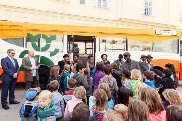 Eine Gruppe Kinder steht vor einem Bus des „Dschungel Wien“ und wird von mehreren Erwachsenen begrüßt.