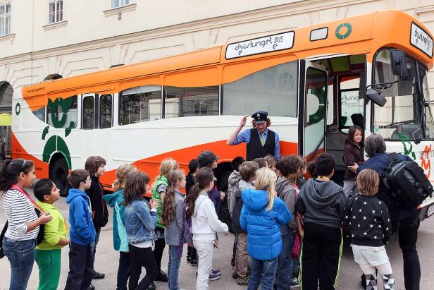 Eine Gruppe Kinder steht vor einem orangefarbenen „Özchunge! BUS“, während ein Busfahrer seinen Hut zieht.