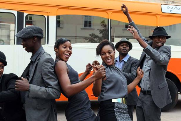 Eine Gruppe junger Leute tanzt und lacht vor einem orangefarbenen Bus.