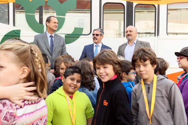 Eine Gruppe von Kindern steht vor einem Bus, hinter ihnen drei Männer in Anzügen.