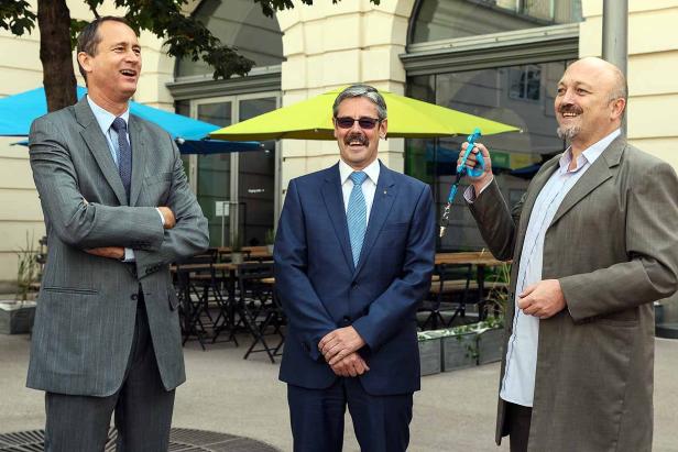 Drei Männer in Anzügen stehen vor einem Café im Freien.