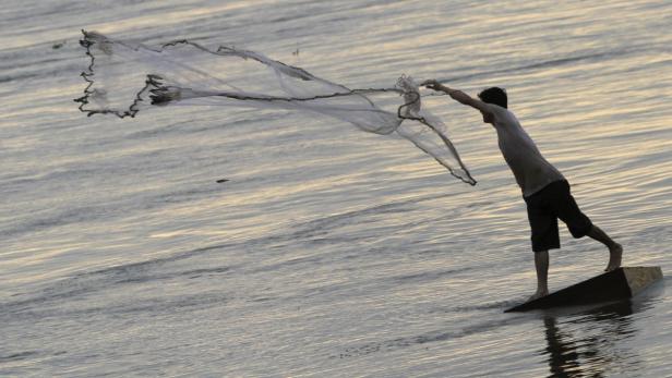 Ein Mann steht auf einem Brett im Wasser und wirft ein Fischernetz aus.