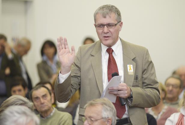 Ein Mann in einem Anzug spricht mit erhobener Hand vor einem Publikum.