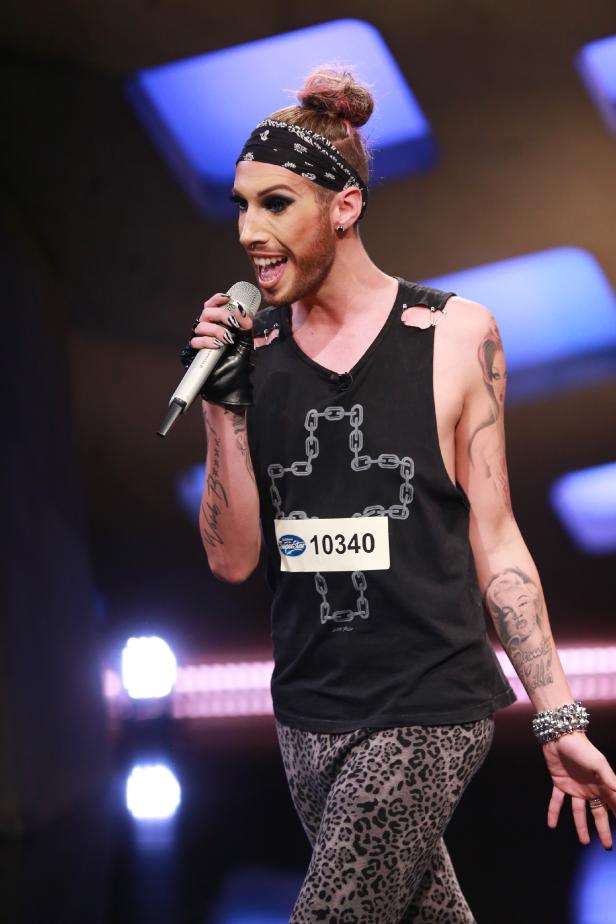 Ein Sänger mit Make-up, Tattoos und einem Stirnband singt in ein Mikrofon.