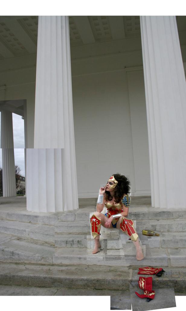 Eine Frau, verkleidet als Wonder Woman, sitzt rauchend auf einer Treppe vor einem Gebäude mit Säulen.