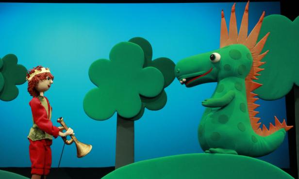 Ein Prinz mit Krone spielt Trompete vor einem grünen Stoffdrachen.