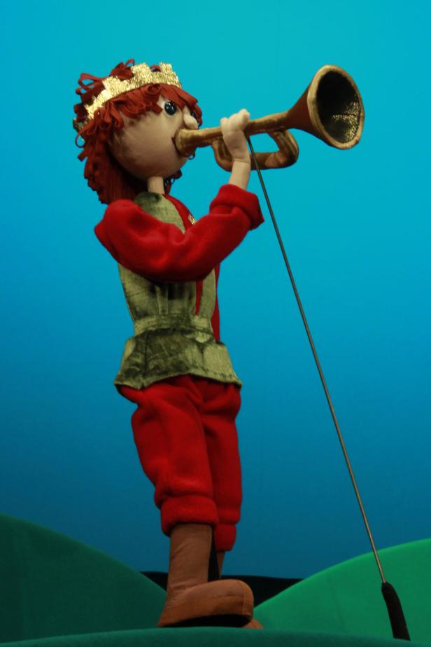Eine Stoffpuppe in königlicher Kleidung spielt auf einer goldenen Trompete.