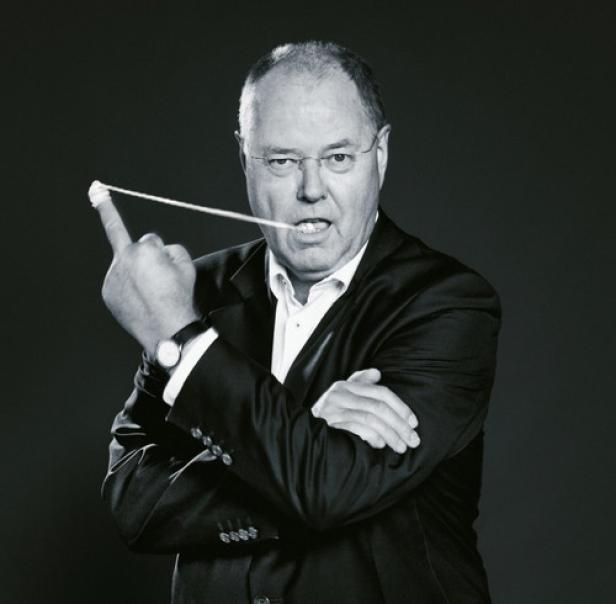 Ein Mann in Anzug und Brille hält ein Gummiband zwischen Finger und Mund.