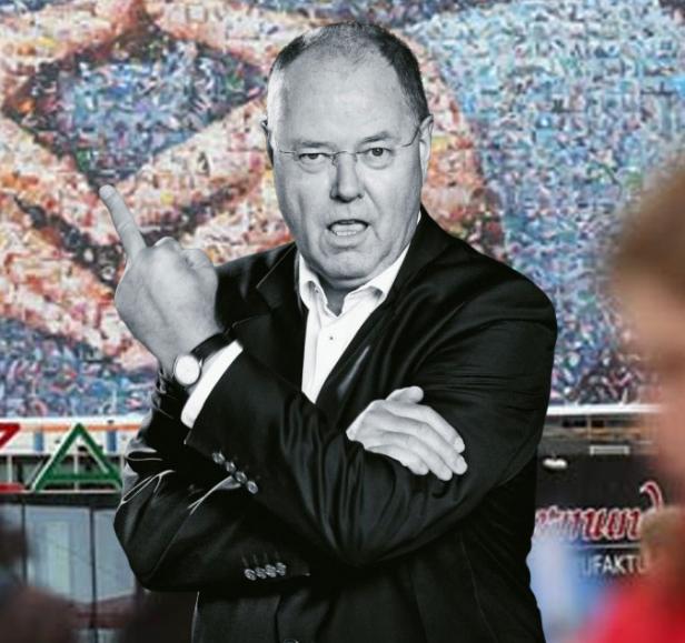 Ein Mann im Anzug zeigt den Mittelfinger vor einem unscharfen Hintergrund.