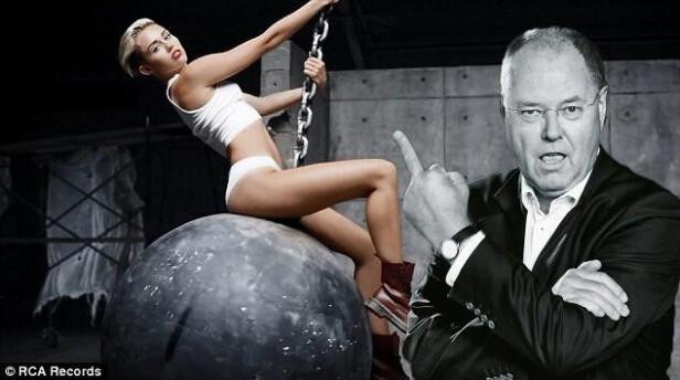 Miley Cyrus posiert auf einer Abrissbirne, während eine Person im Anzug den Mittelfinger zeigt.