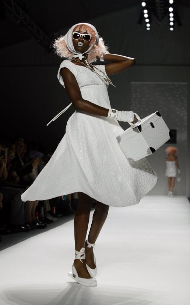 Eine dunkelhäutige Frau mit rosa Perücke und weißem Kleid auf einem Laufsteg.