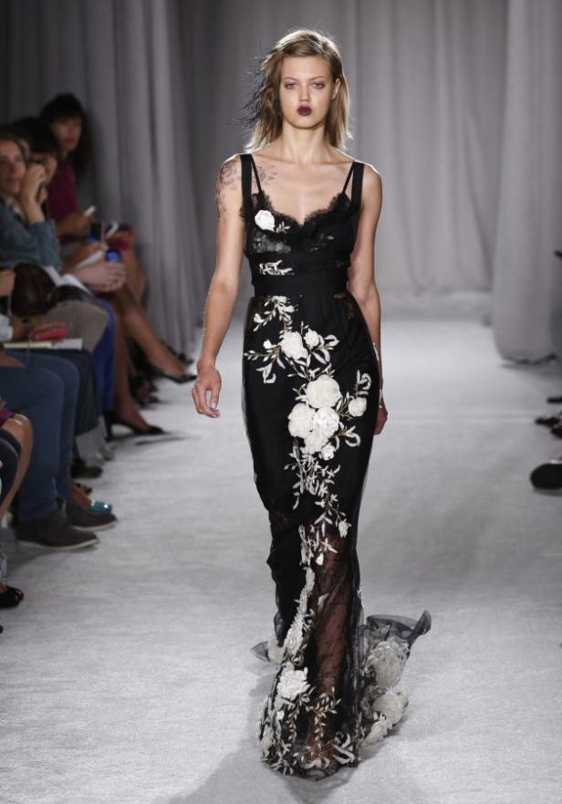 Ein Model präsentiert ein schwarzes Abendkleid mit weißen Blumenapplikationen auf dem Laufsteg.