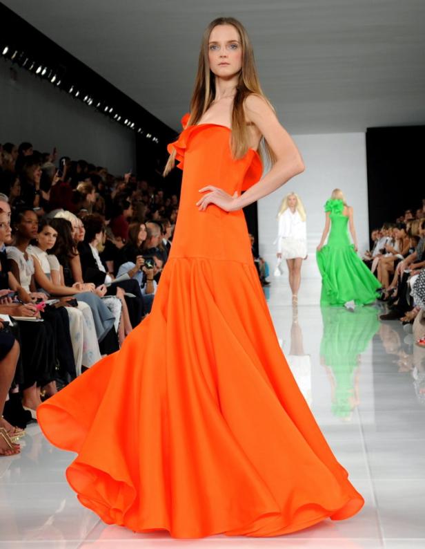 Ein Model präsentiert ein orangefarbenes, bodenlanges Kleid auf dem Laufsteg.