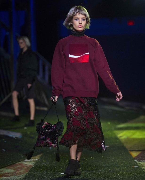 Ein Model auf dem Laufsteg, gekleidet in einem burgunderroten Pullover und einem gemusterten Rock.