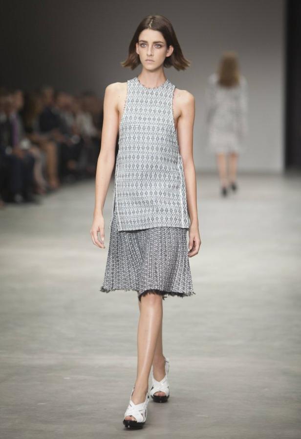 Eine Frau präsentiert ein ärmelloses Kleid und weiße Schuhe auf einem Laufsteg.