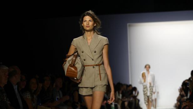 Eine Frau präsentiert ein beiges Hosenanzug und eine braune Handtasche auf dem Laufsteg.