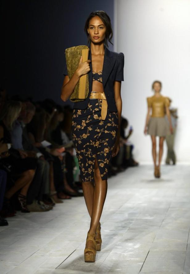 Eine Frau präsentiert ein blaues, florales Ensemble mit Blazer und Clutch auf dem Laufsteg.