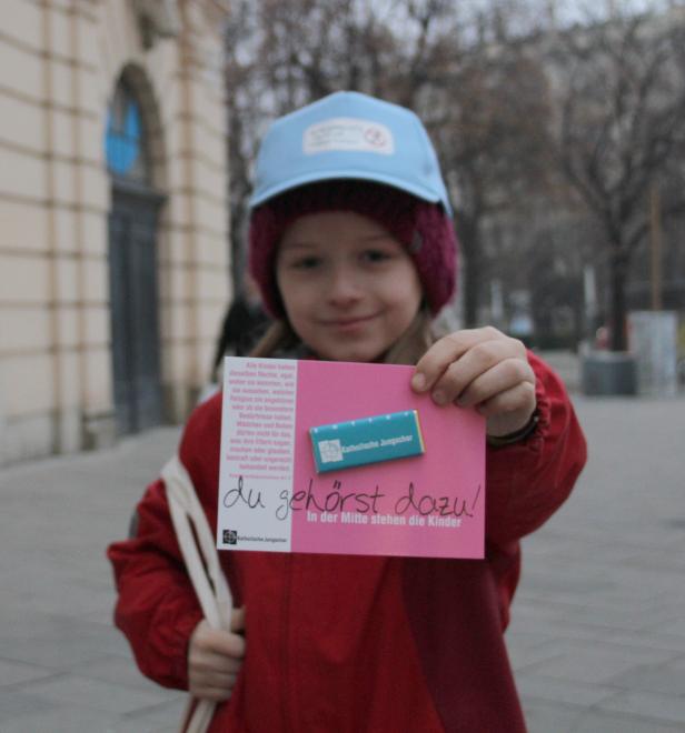 Ein Mädchen hält ein Schild der Katholischen Jungschar mit der Aufschrift „du gehörst dazu!“.