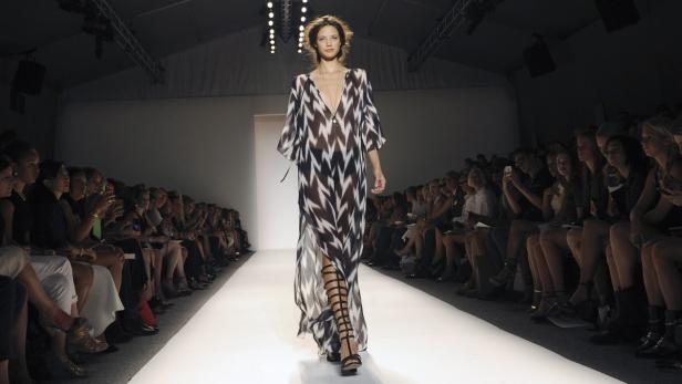 Eine Frau präsentiert ein langes, gemustertes Kleid und Gladiatorensandalen auf einem Laufsteg.