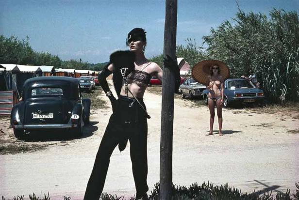 Eine androgyn gekleidete Person posiert vor einem Oldtimer und einer Frau mit Sonnenschirm am Strand.