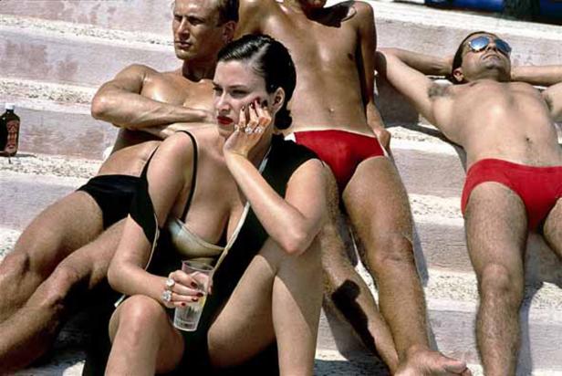 Eine Frau sitzt mit einem Drink auf Stufen, umgeben von sonnenbadenden Männern.