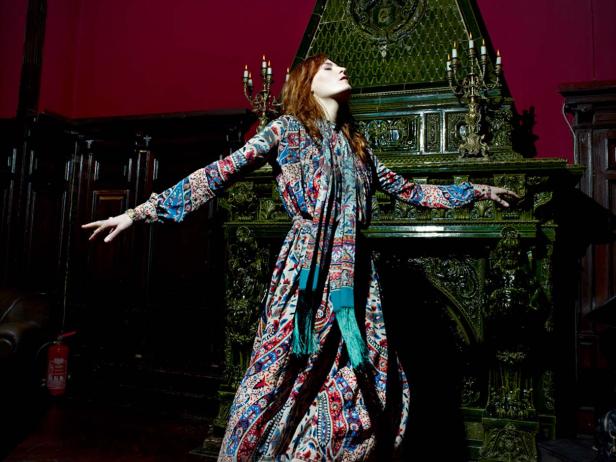 Eine Frau in einem bunten Kleid steht vor einem verzierten, grünen Kamin.