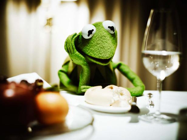 Kermit der Frosch sitzt an einem gedeckten Tisch und scheint nachdenklich.