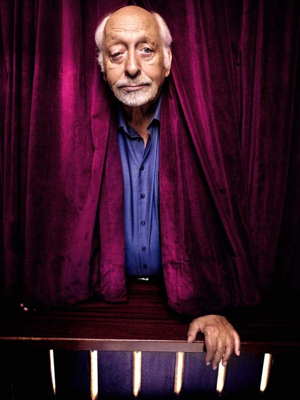 Ein älterer Mann mit Bart schaut hinter einem roten Vorhang hervor.