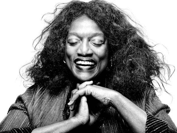 Schwarzweißporträt einer lachenden Frau mit lockigem Haar und verschränkten Händen.