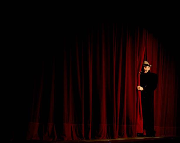 Ein Mann mit Hut lugt hinter einem roten Theatervorhang hervor.