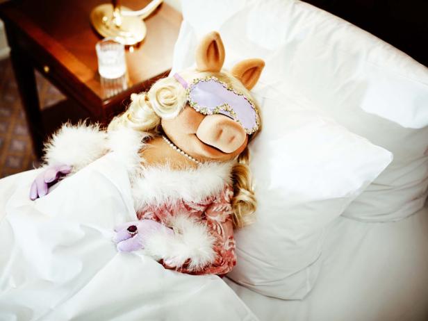 Miss Piggy schläft mit Schlafmaske, Handschuhen und Schmuck in einem Bett.