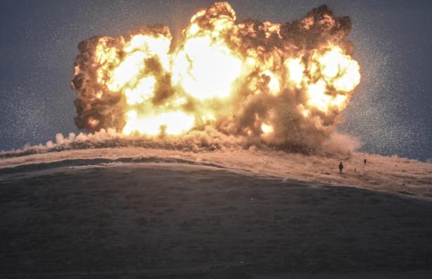Eine große Explosion auf einem Hügel, mit zwei Personen im Hintergrund.