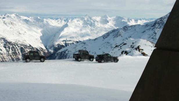 Drei Geländewagen fahren auf einer schneebedeckten Fläche vor einer Bergkulisse.