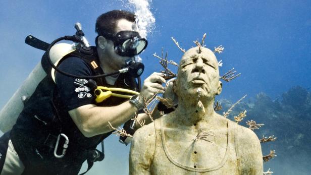Ein Taucher interagiert unter Wasser mit einer Statue, die mit Korallen bewachsen ist.