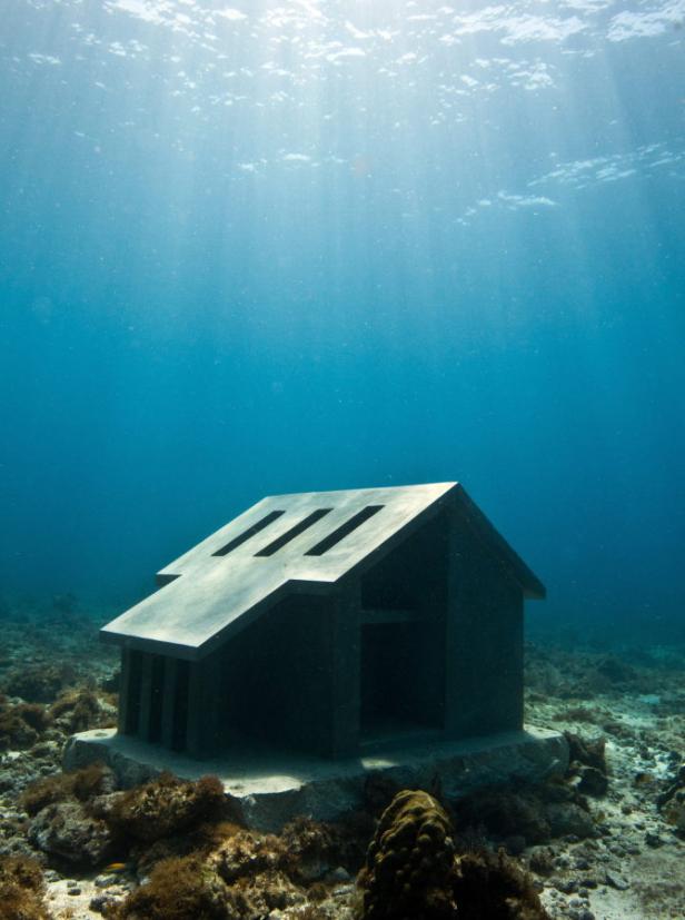 Ein skulpturales Haus steht unter Wasser auf einem bewachsenen Meeresgrund.