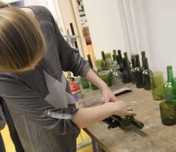 Eine Frau schneidet eine grüne Glasflasche auf einem Tisch mit vielen Flaschen.